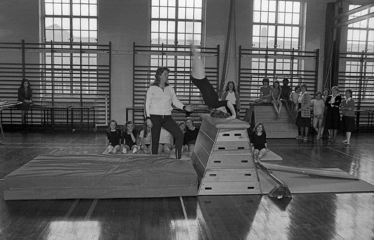 Black-and-white photo of a girl vaulting head-first over a wooden vaulting box onto a crash mat while a woman teacher in a tracksuit stands ready; girls in leotards sit on mats and others watch from wall bars beneath tall windows.