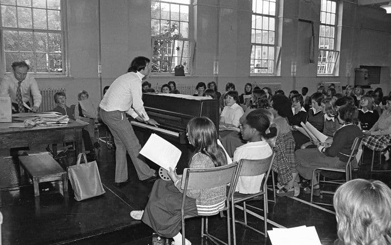 Black-and-white photo of a crowded school hall: a teacher in flared checked trousers plays an upright piano on a low stage while rows of pupils holding song sheets sing, and another teacher sorts papers at a table on the left.