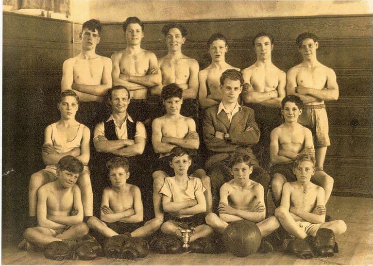 Sepia group photograph of a boys’ and young men’s boxing team in three rows in a wood-panelled hall, most bare-chested with arms folded, two men in waistcoat and jacket in the middle row, and boxing gloves, a football and a small trophy cup on the floor in front.