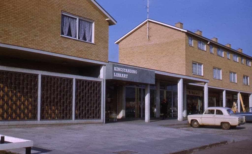 1960s: Colour photograph of the modern Kingstanding Library, with a “Kingstanding Library” sign over a glazed entrance and honeycomb screen walls, set into a parade of yellow-brick shops with flats above; a white Ford Popular is parked at the kerb.