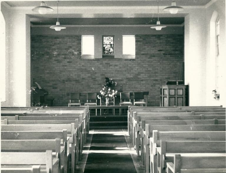 Black-and-white photo of a plain modern chapel interior: rows of light wooden pews either side of a carpeted aisle lead to a raised platform with chairs and a vase of flowers, a brick end wall with three small windows, and a pulpit on the right.