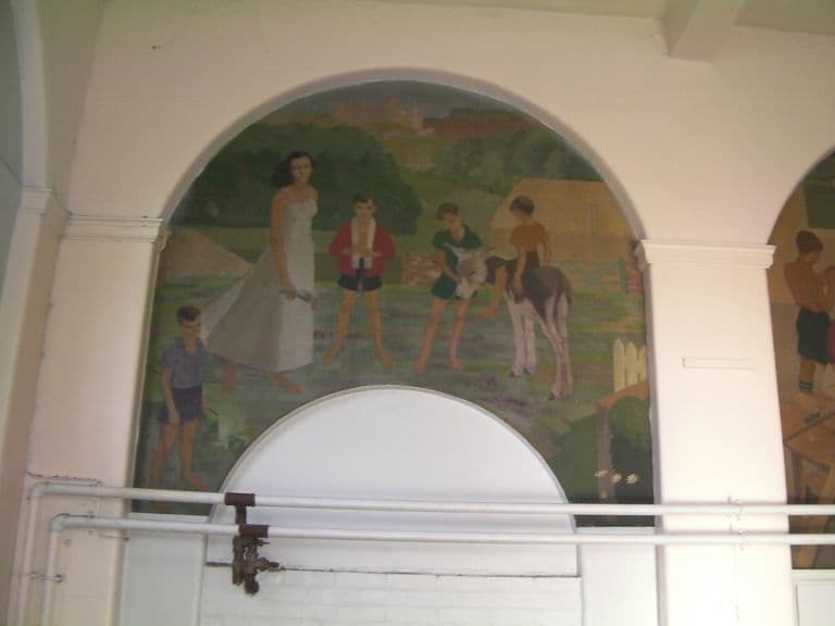An arched mural of a young woman in a long white dress walking barefoot with children in a meadow, one child riding a donkey, with a castle on a hill in the background.