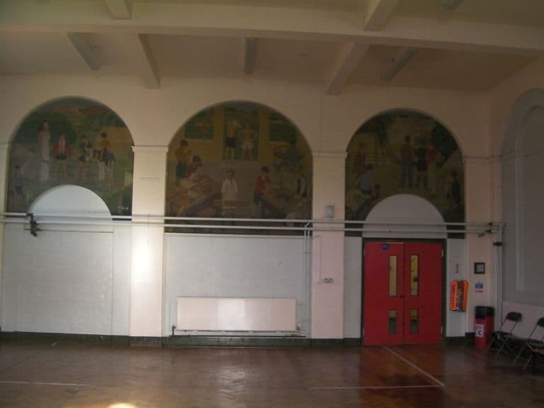 A hall with a parquet floor and three large arched mural panels high on the wall, showing children and young people in the countryside, at workbenches and in a field; heating pipes run across below and a red double door stands at the right.