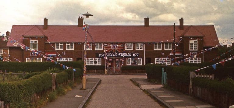 Colour photograph of a terrace of red-brick council houses hung with red, white and blue bunting and Union Jacks, with a banner reading “1952 Silver Jubilee 1977” across the front and a striped lamp post in the foreground.