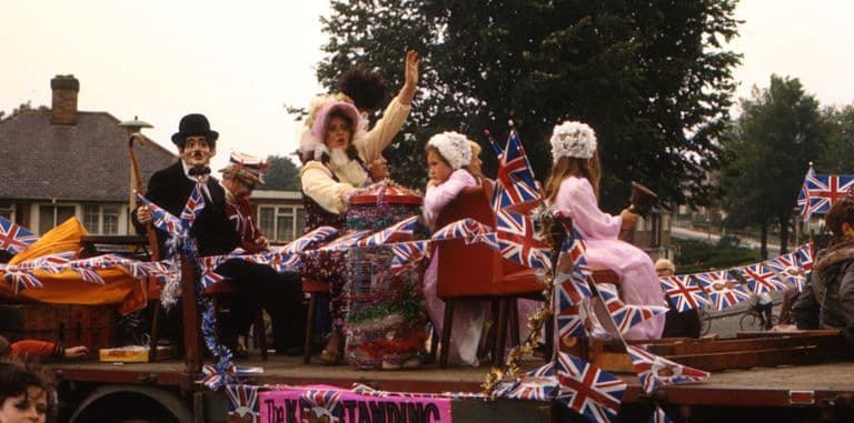 Colour photograph of a lorry float draped in Union Jack bunting carrying people in costume, including a Charlie Chaplin figure, a woman in a bonnet waving and girls in pink gowns, with a “The Kingstanding …” banner on the side.