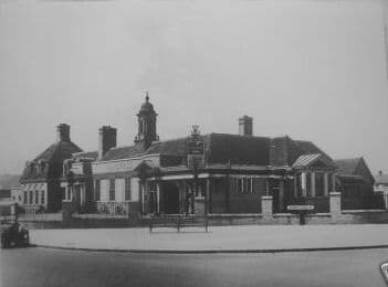 A small photograph of the College Arms: a large brick pub with a small domed cupola, tall chimneys, a pillared entrance and a hanging sign, set behind a low wall with gate piers, a road sign and an old car on the left.