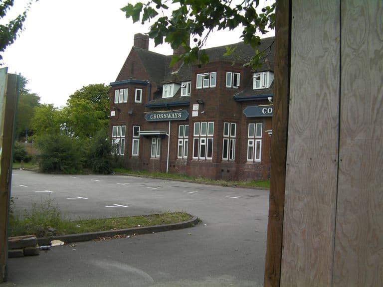A large red-brick pub with tall chimneys, bay windows and a “Crossways” sign, seen through a gap in wooden hoarding across an empty car park with overgrown shrubs.
