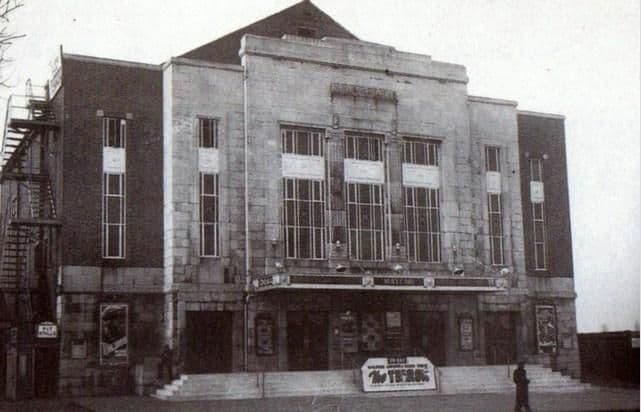 The stone-faced front of the Mayfair cinema: a tall central block with three large windows, brick side wings, a canopy lettered “Mayfair”, a steel fire escape on the left, film posters by the doors, a board on the steps and one man walking past.