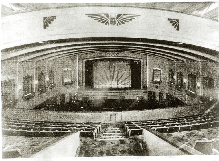 The auditorium of the Mayfair cinema seen from the back of the circle: curved balcony fronts, a large winged “M” emblem above the proscenium, a sunburst-patterned stage curtain, wall lamps in decorative frames, rows of empty seats and carpeted steps with an “M” pattern.