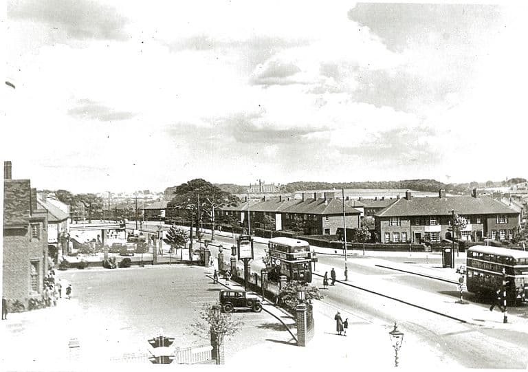 A raised view along College Road: the Crossways pub’s gate piers, hanging sign and forecourt on the left with a parked car, two double-decker buses and pedestrians on the road, rows of new brick houses across the junction, and open country and a large building on the skyline.