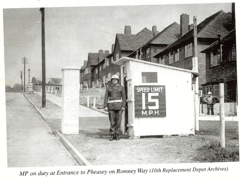 A scan of a book page: an American military policeman in a white helmet and belt stands by a white sentry box with a “Speed limit 15 m.p.h.” sign, at a gateway beside a row of houses on a sloping road; another soldier stands behind a fence. The printed caption reads “MP on duty at Entrance to Pheasey on Romney Way (10th Replacement Depot Archives)”.
