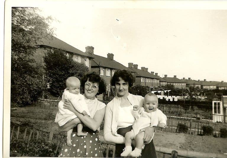 Black-and-white snapshot of two smiling young women each holding a baby in knitted clothes, standing by a garden fence; behind them a long row of brick council houses with tall chimneys, and washing on a line in the back gardens.