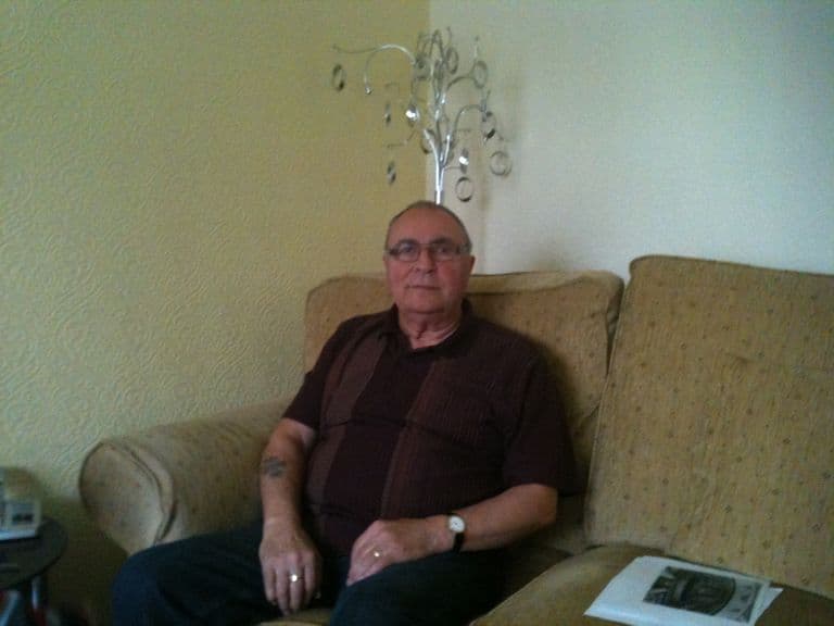 An older man with glasses in a dark striped shirt, seated on a sofa with papers beside him.