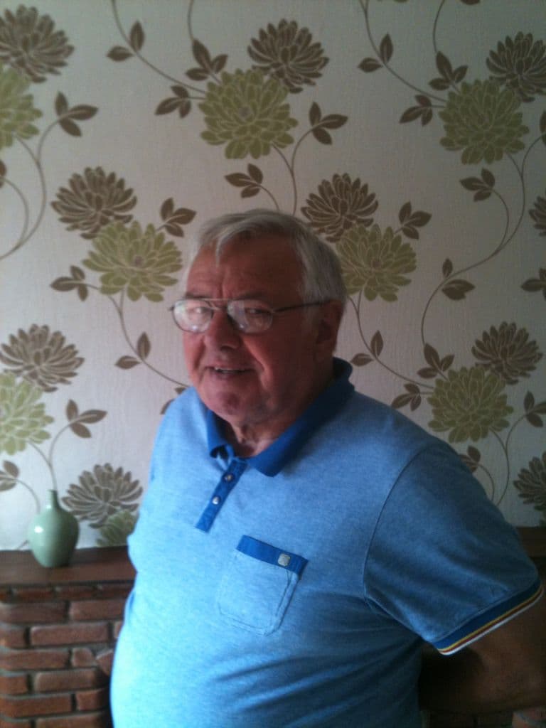 An older man with white hair and glasses in a blue polo shirt, standing in front of floral wallpaper.