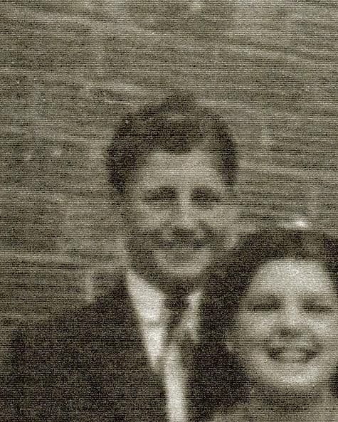 A grainy old photograph of a smiling young man with dark wavy hair in a suit and tie, with a smiling young woman partly in frame, against a brick wall.