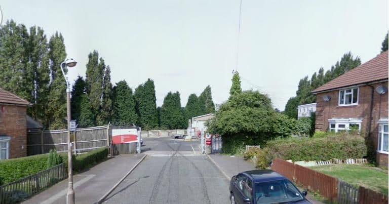 A street view along Ealing Grove to the gates of the council depot at its end, with a red sign on the fence, tall poplar trees behind, a lamp post, hedges and a car parked outside a house on the right.