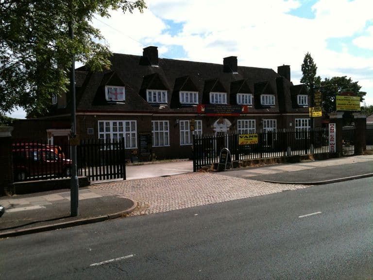 The long two-storey Hare and Hounds building with dormer windows and tall chimneys behind black railings, its forecourt in use as a valeting centre and hand car wash, with yellow and red signs along the fence.