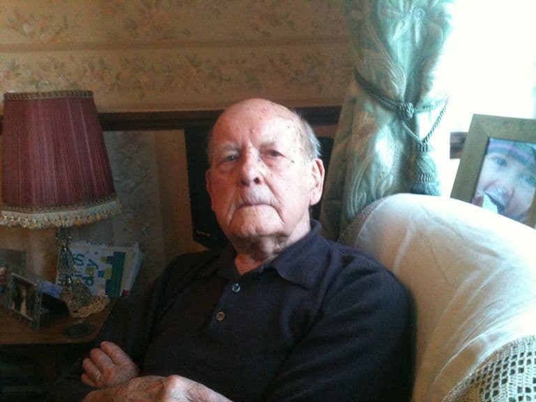 An older man in a dark polo shirt seated in an armchair beside a lamp and birthday cards.