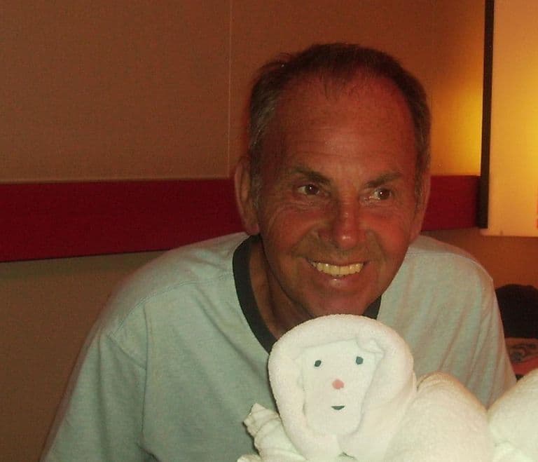 A smiling man in a pale T-shirt seated at a table, with a folded towel animal in front of him.