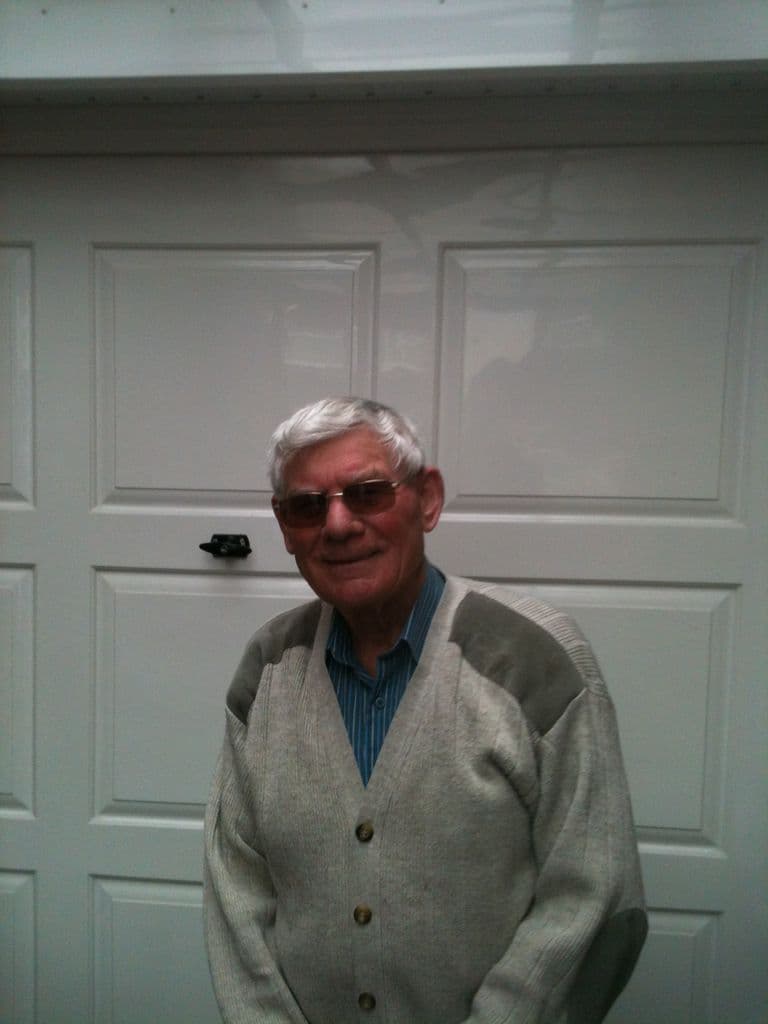 An older man with white hair and tinted glasses in a grey cardigan, standing in front of a white garage door.