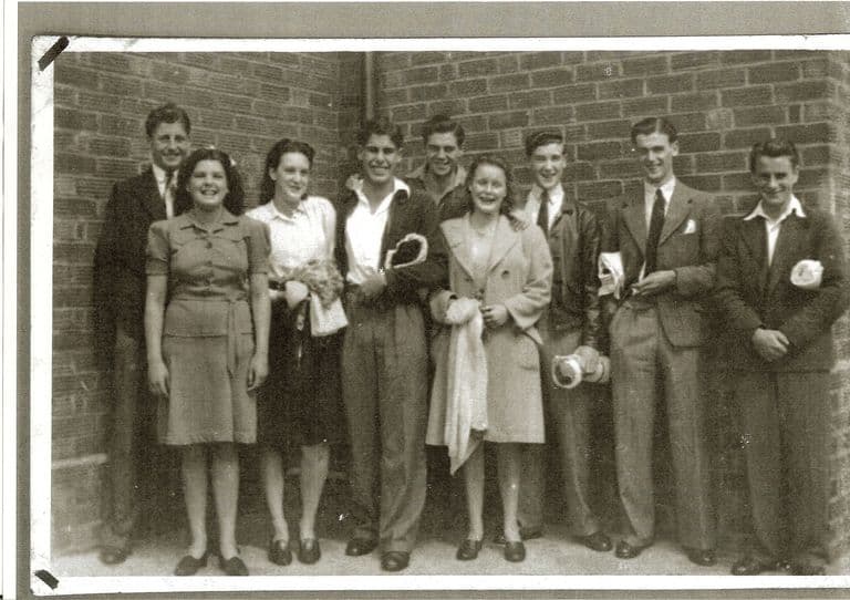 Black-and-white snapshot of nine laughing young men and women in 1940s clothes standing against a brick wall, several holding rolled towels and swimming things.