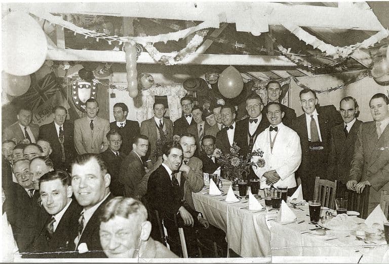 Black-and-white photograph of a crowded club dinner: rows of men in suits and bow ties at long tables laid with white cloths, pints of beer and flowers, under paper streamers and balloons; a British Legion banner and a shield hang on the wall.