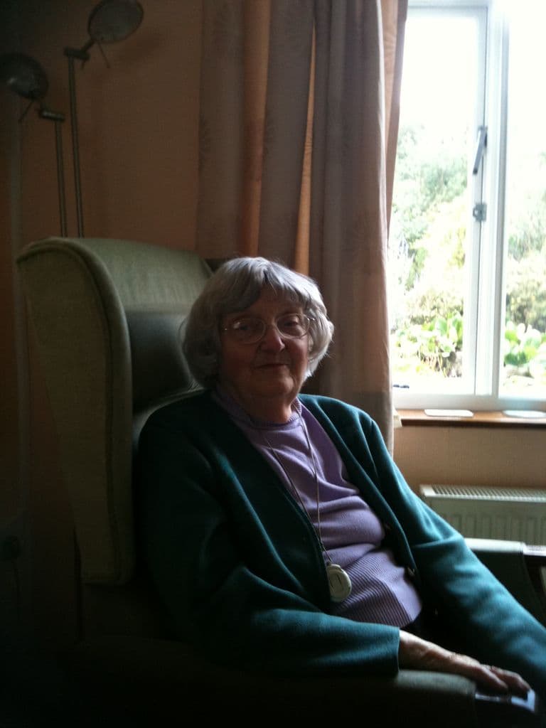 An older woman with grey hair and glasses in a green cardigan, seated in a wing chair by a window.