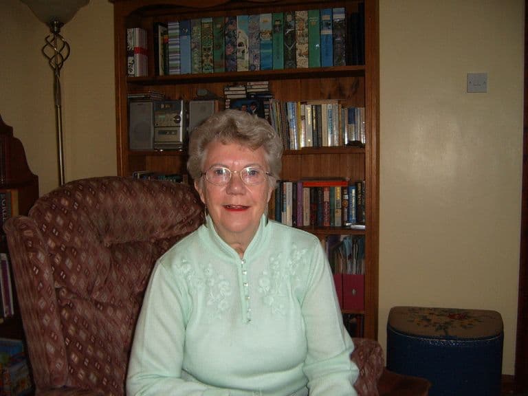 An older woman with grey hair and glasses in a pale green jumper, seated in an armchair in front of a bookcase.