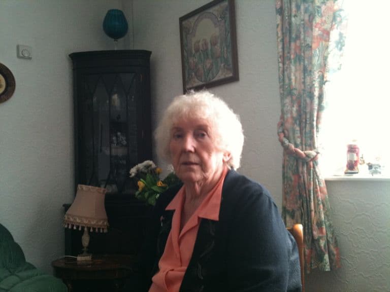 An older woman with white hair, in a navy cardigan and coral blouse, seated in a living room with a corner cabinet and flowers.