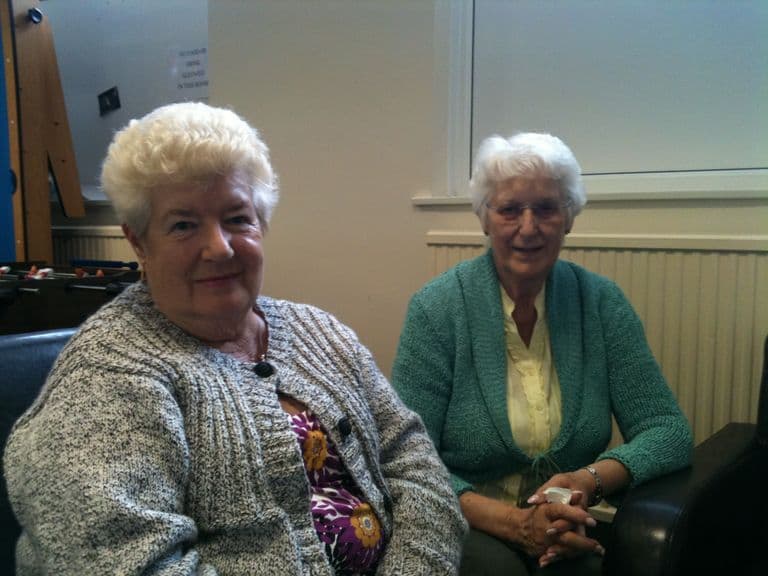 Two older women with white hair seated side by side in a community room, one in a grey knitted cardigan and one in a green cardigan.