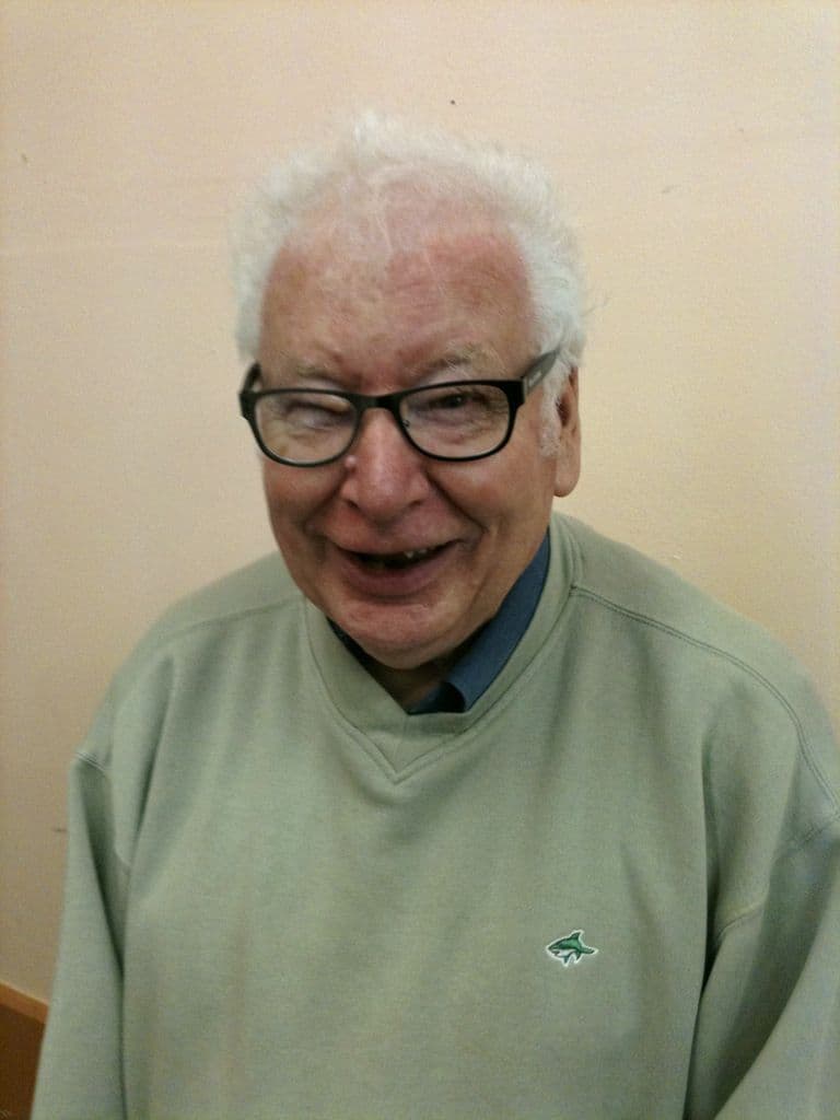 Head-and-shoulders portrait of an older man with white hair and dark-rimmed glasses, laughing, in a green sweatshirt against a cream wall.