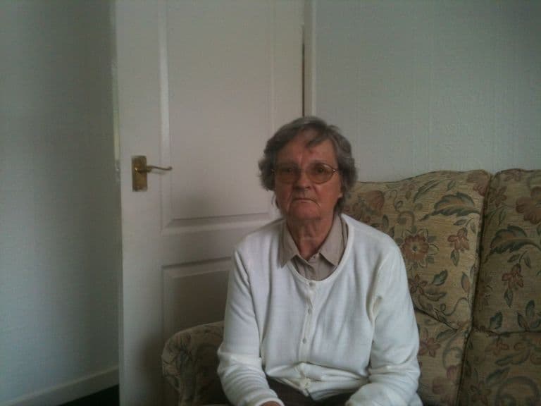 An older woman with grey hair and glasses in a white cardigan, seated on a floral sofa.