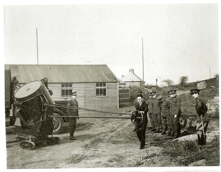 A man in a dark suit turns a control wheel linked by long rods to a large searchlight on a tracked mounting, watched by about ten soldiers in Territorial Army uniform and an officer with a swagger stick, beside a wooden hut, a lorry and a house.