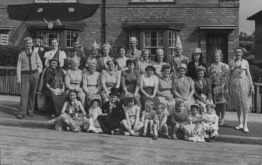 Around forty women and children, many in paper hats, crowns and fancy dress – including grass skirts and a top hat – pose in rows on the pavement in front of brick houses; a banner hangs above the bay windows.