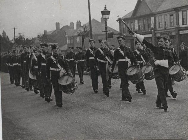 Air Training Corps cadets in forage caps and white belts march down a road behind drummers and a drum major with a raised mace; shops and a street lamp stand behind them.