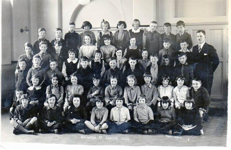Class photograph of about forty boys and girls in four rows in a school hall, with a moustached male teacher standing at the right; written along the bottom of the print: “Dulwich Rd Junior 1932” and “Photo V. Cook”.