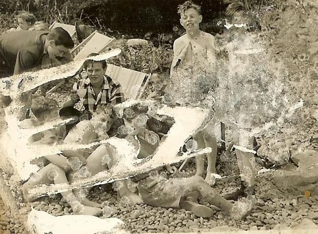 A badly damaged print of boys and young men relaxing on a pebble beach with deckchairs; large areas of the emulsion have flaked away across the centre and right.