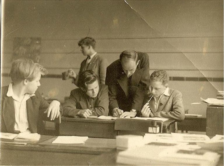 A creased sepia classroom photo of a tall teacher in a suit leaning over a desk to help a boy with his work while other teenage boys sit and stand around him; books and papers are piled in the foreground.