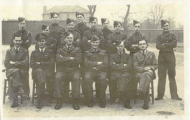 Group portrait of about seventeen Air Training Corps cadets in forage caps standing behind six seated officers and instructors, most with arms folded, in front of a brick wall and a house.