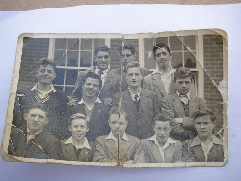 A cracked and creased black-and-white print of thirteen teenage boys in jackets and ties in three rows in front of a brick school building with sash windows.