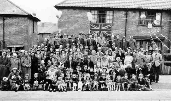 A large group of residents – several rows of adults and children seated on the pavement in front – gathered in the street for the coronation, with a Union flag on a house, bunting and balloons.