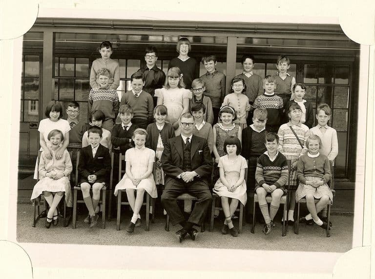 Class of about thirty junior-school children in four rows on the playground, a male teacher in glasses and a three-piece suit seated in the centre of the front row, in front of a veranda with glazed doors.