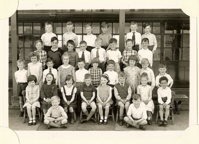 Class of about forty infants in five rows, two boys sitting cross-legged on a mat in front, in front of a veranda with glazed doors; no teacher appears.