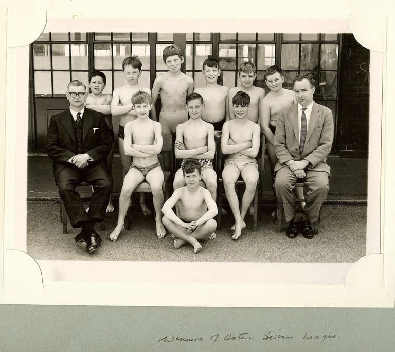 A school team photograph of eleven boys in swimming trunks with two male teachers in suits, in front of the school’s glazed doors, mounted in an album with the handwritten caption “Winners of Aston Swim League”.