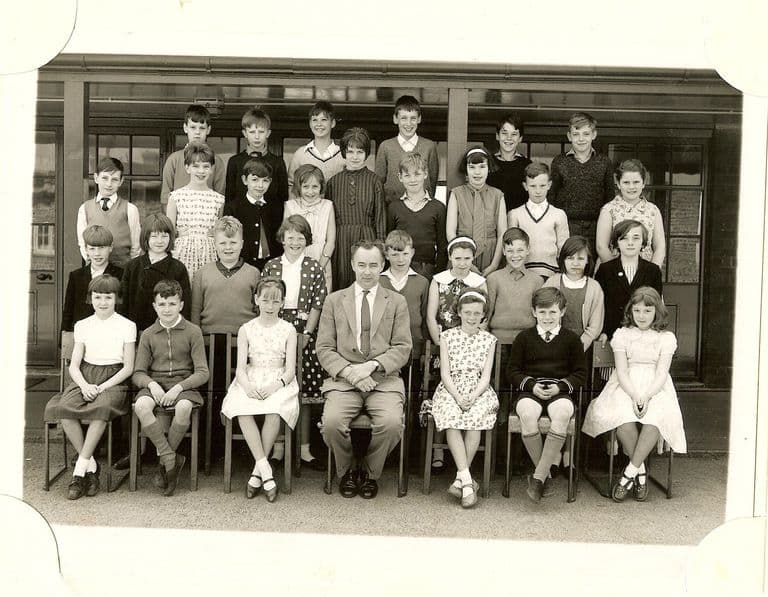 Class of about thirty-three junior-school children in four rows, a male teacher in a light jacket and striped tie seated in the centre of the front row, in front of glazed classroom doors.