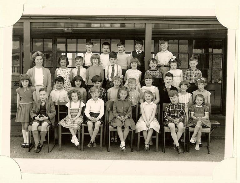 Class of about thirty-four young children in four rows, a smiling woman teacher in a cardigan standing at the left, in front of a veranda with glazed doors.