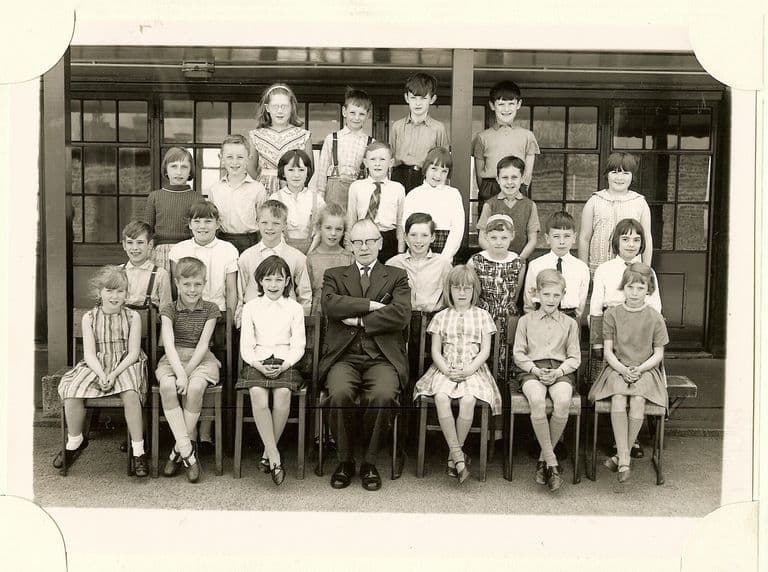 Class of about twenty-seven junior-school children in four rows, an older male teacher in glasses seated in the centre with his arms folded, in front of glazed classroom doors.
