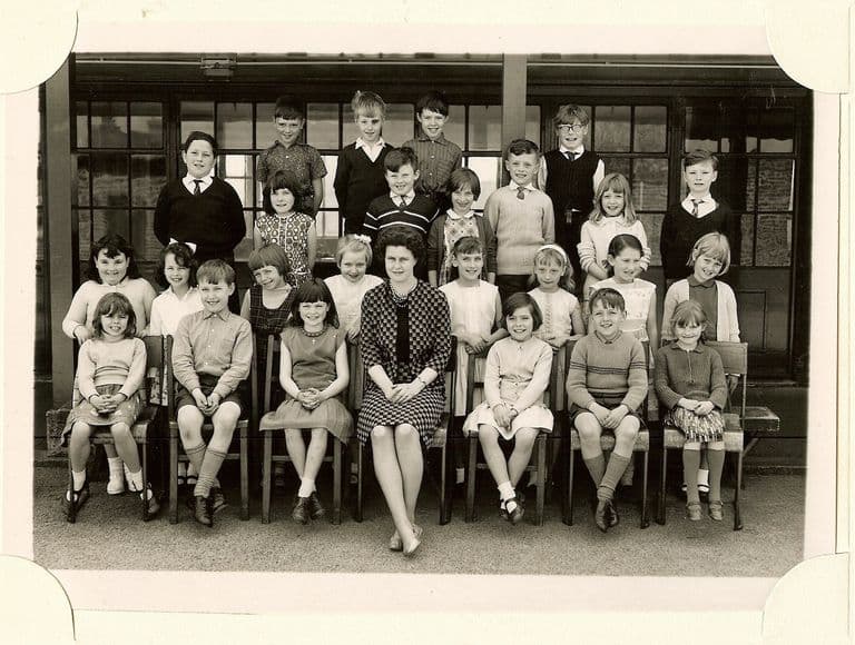 Class of about thirty-three children in four rows, a woman teacher in a checked dress seated in the centre of the front row, in front of a veranda with glazed doors.