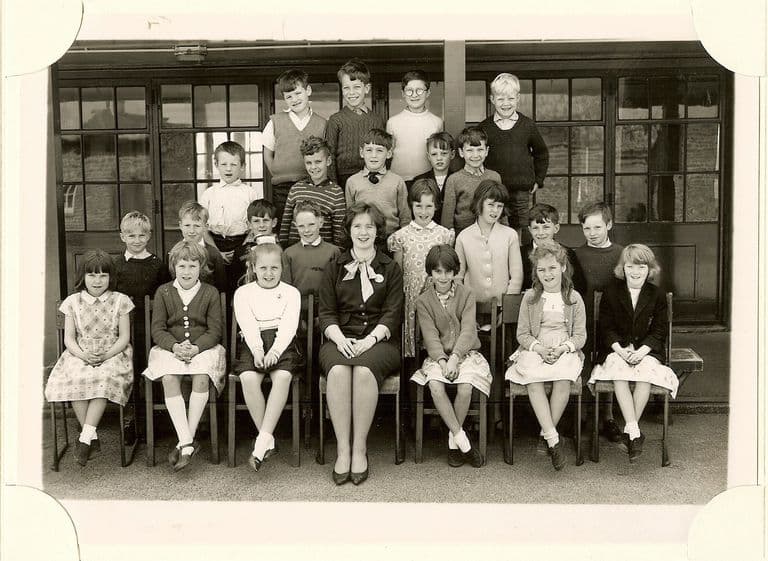 Class of about thirty-three children in four rows, a woman teacher in a dark dress with a bow at the neck seated in the centre of the front row.