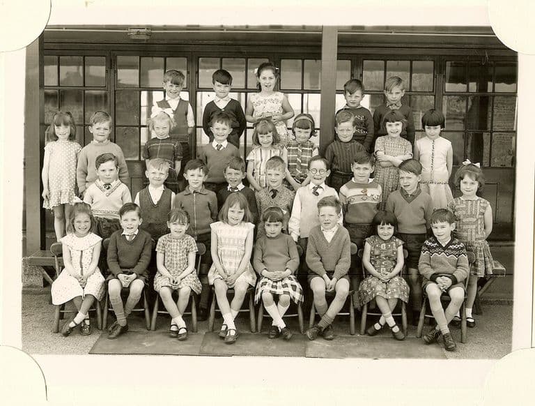 Class of about thirty-eight infants in four rows, the front row on small chairs set on mats, in front of a veranda with glazed doors; no teacher appears.